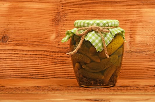 One glass jar of homemade pickled cucumbers with green checkered textile top decoration at brown painted vintage wooden surface