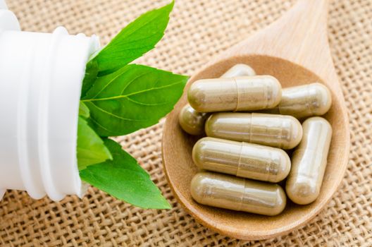 Herb capsule spilling out of a bottle in wooden spoon with green leaf on sack background.
