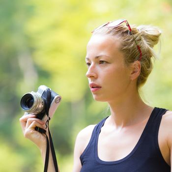 Portrait of eautiful blonde caucasian girl wearing sporty black sleeveless t-shirt, outdoors in nature, holding vintage camera in her hand. Square composition.