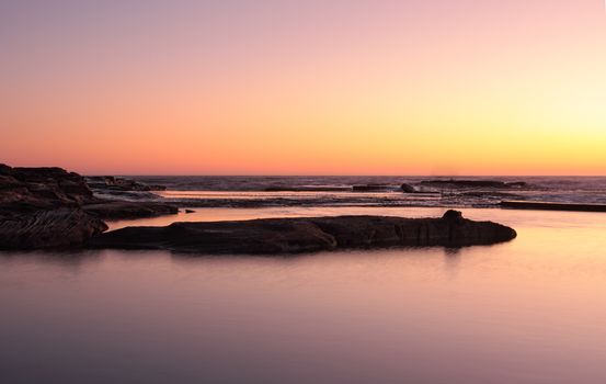 The golden glow of the suns rays breaking forth colour the sky just before sunrise.  Simple views and almost silhouettes across the rock pool to the ocean. Simple image suitable as a background image or your message