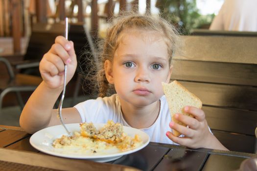 The four-year girl sits at a table and eat a main dish with bread in an outdoor cafe