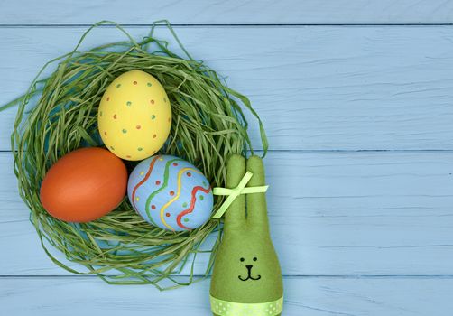 Easter eggs and rabbit. Happy bunny handmade and hand painted multicolored decorated eggs on green straw nest, blue wooden background, copyspace. Unusual creative holiday greeting card 