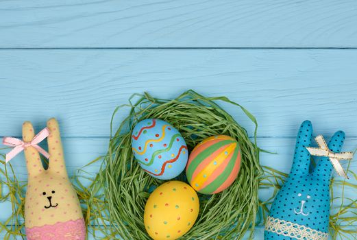 Easter eggs and rabbits. Happy bunny handmade and hand painted multicolored decorated eggs on green straw nest, blue wooden background, copyspace. Unusual creative holiday greeting card 