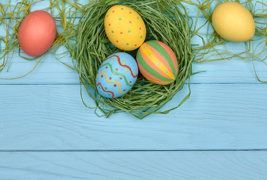 Easter eggs background. Hand painted multicolored decorated eggs on green straw nest, blue wood, copyspace. Unusual creative holiday greeting card 