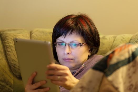 housewife middle age woman without makeup reading news on tablet evening at home on sofa