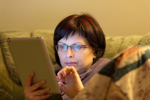 housewife middle age woman without makeup reading news on tablet evening at home on sofa
