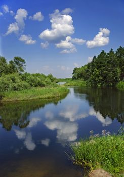 Summer landscape with calm river, mixed forest, cloudy sky and the reflection in the water.