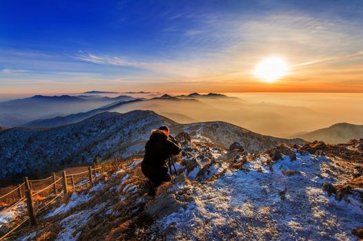 Professional photographer takes photos with camera on tripod on rocky peak at sunset.