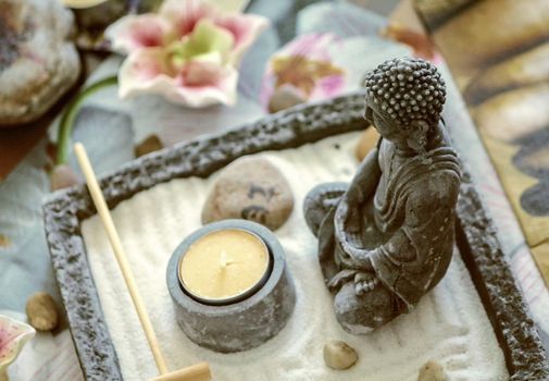 Small statue of Buddha meditating in sand next stones and lotus flowers