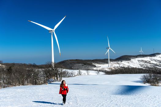 Beautiful girl walking in winter landscape of sky and winter road with snow and red dress and wind turbine.