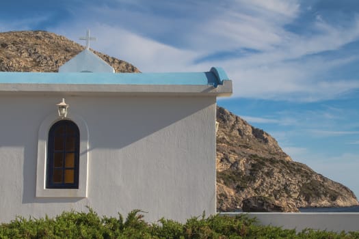 White traditional greek church surrounded by mountains from one side and a sea from another side. Blue sky with many white clouds.