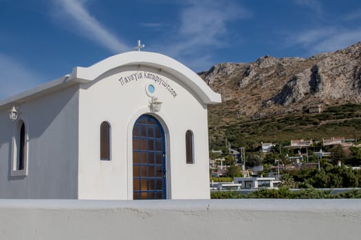 White traditional greek church surrounded by mountains from one side and a sea from another side. Blue sky with many white clouds.