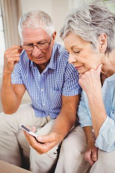 Sad senior couple sitting on sofa and looking at mobile phone in living room