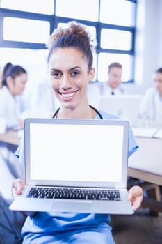 Portrait of female doctor showing laptop and other doctors discussing behind in conference room