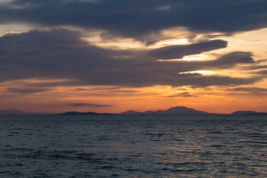 Dark water of the Aegean sea, with reflection from the sunlight during sunset. Cloudy sky. Mountains in the background.