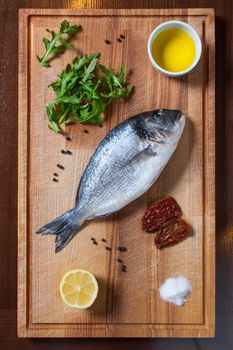 Fresh uncooked dorado or sea bream fish with tomatoes, lemon, arugula, olive oil and spices on rustic wooden cutting board, top view