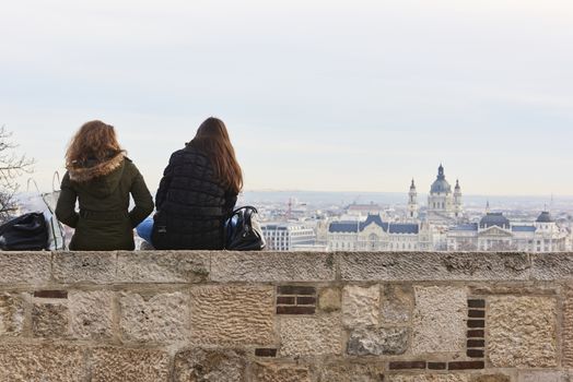 BUDAPEST, HUNGARY - FEBRUARY 02: Two girls sitting on wall enjoying the view from Buda Castle, with Gresham Palace and Saint Stephen's Basilica in the background. February 02, 2016 in Budapest.