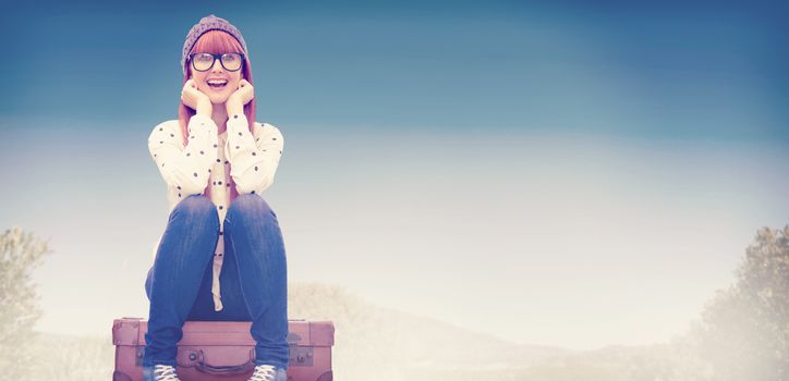 Smiling hipster woman sitting on suitcase  against mountain trail