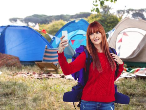 Smiling hipster woman with a travel bag taking selfie against empty campsite at music festival
