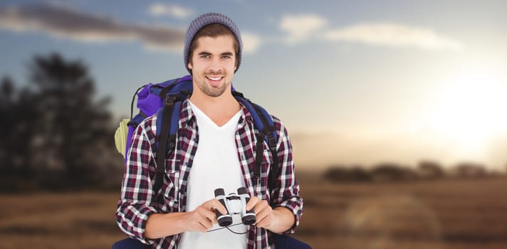 Happy hipster wearing backpack holding binoculars against landscape of the countryside