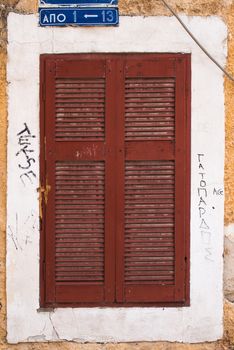 Window covered by a closed dark red shutter. White frame around the window. Small frame made by yellow facade painting.
