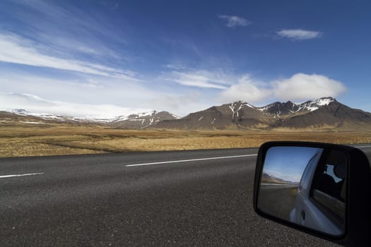 Horizontal photo of a car mirror on the side of a paved road in Iceland with mountains and blue sky in the background