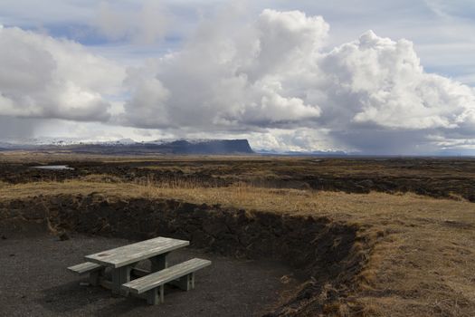 Horizontal panorama of a wooden resting bench with mountains covered in snow and dramatic clouds above 