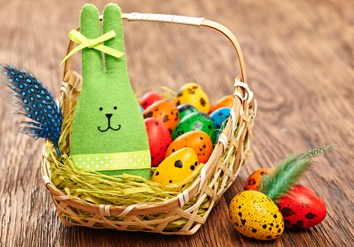 Easter rabbit and eggs in basket. Happy bunny handmade and hand painted multicolored quail with straw and feathers, on wooden background. Unusual creative holiday greeting card 