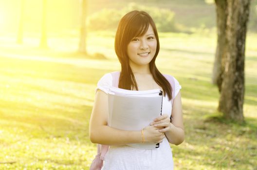 Young Asian teen student standing on campus lawn, holding books and smiling.