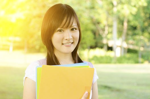 Young Asian college student standing on campus lawn, holding file folder and smiling.