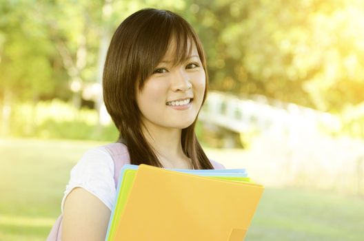 Young Asian university student standing on campus lawn, holding file folder and smiling.