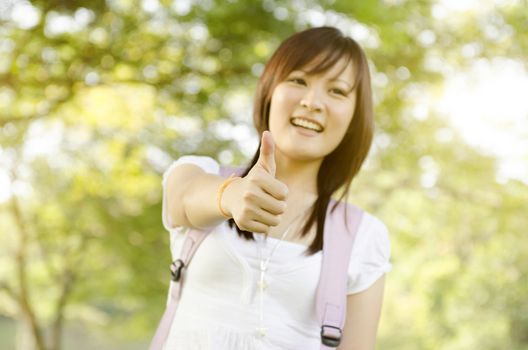 Young Asian college girl student standing on campus lawn, giving thumb up and smiling.