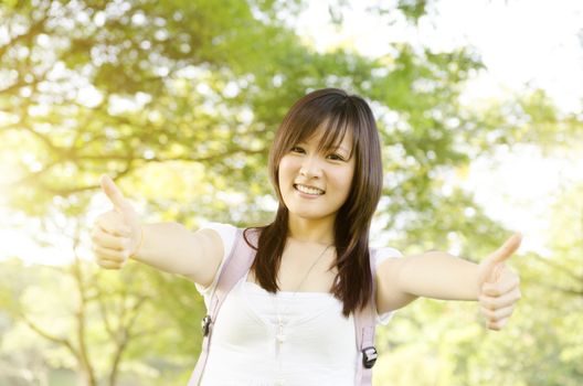 Young Asian college girl student standing on campus lawn, showing thumbs up and smiling.