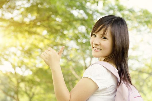 Young Asian college girl student standing on campus lawn, showing thumb up and smiling.