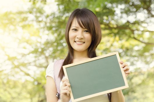 Young Asian college girl student standing on campus lawn, holding a blank chalkboard and smiling.