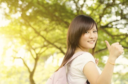 Young Asian university girl student standing on campus lawn, giving thumb up and smiling.