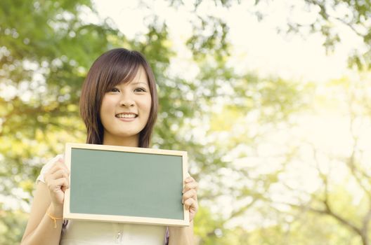 Young Asian university girl student standing on campus lawn, holding a blank blackboard and smiling.