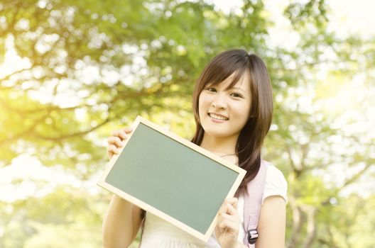 Young Asian university girl student standing on campus lawn, holding a blank chalkboard and smiling.