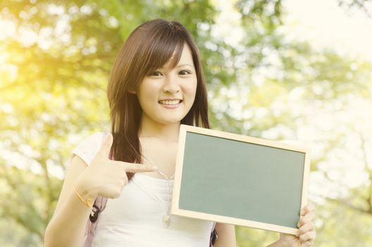 Young Asian college girl student standing on campus lawn, hand pointing to a blank chalkboard and smiling.