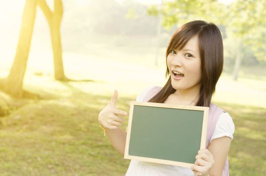 Young Asian teen girl student standing on campus lawn, hands pointing to a blank chalkboard and smiling.