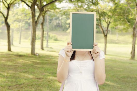 Young Asian college girl student standing on campus lawn, hands holding a blank chalkboard and covered face.