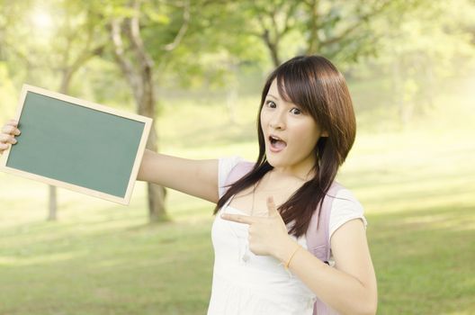 Young Asian college girl student standing on campus lawn, hands holding a blank chalkboard with shocked expression.