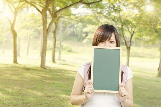 Young Asian college girl student standing on campus lawn, hands holding a blank chalkboard and covered mouth.