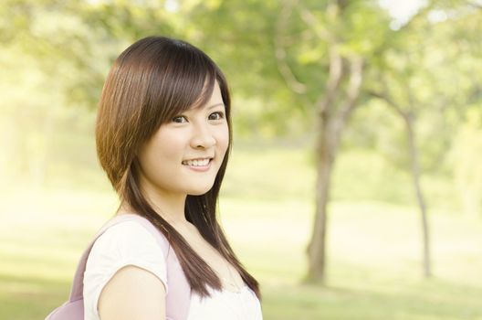 Young female Asian teen student standing on campus lawn, with backpack and smiling.