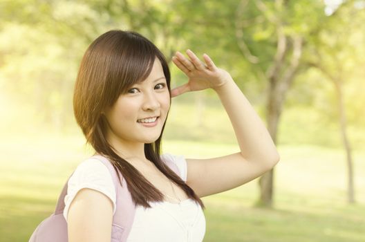 Young Asian college girl student standing on campus lawn, hand shielding and smiling.