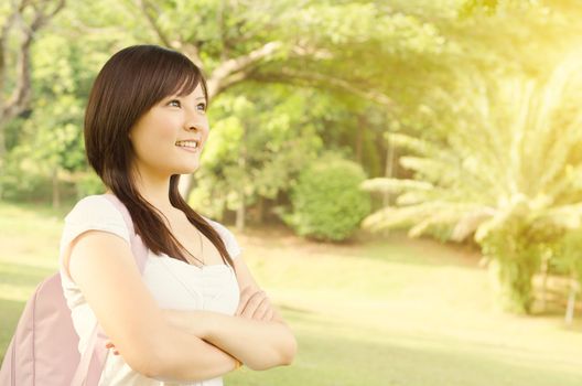 Young female Asian university student standing on campus lawn, with backpack looking away  and smiling.