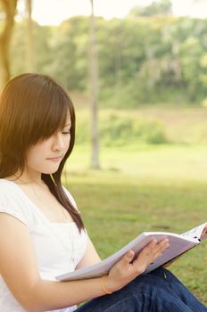 Young Asian college girl student sitting on campus lawn and reading book.