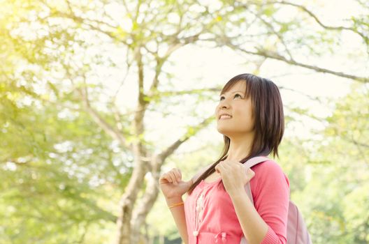 Young Asian college girl student standing on campus lawn, with backpack looking away and smiling.