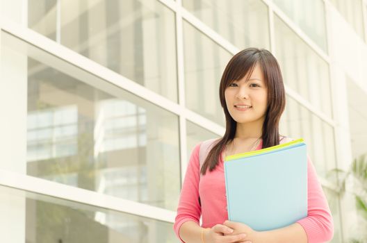 Young Asian college student standing outside campus building, holding file folder and smiling.