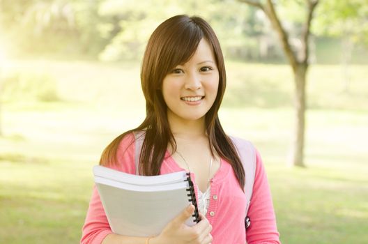 Young Asian university student standing on campus lawn, holding books and smiling.